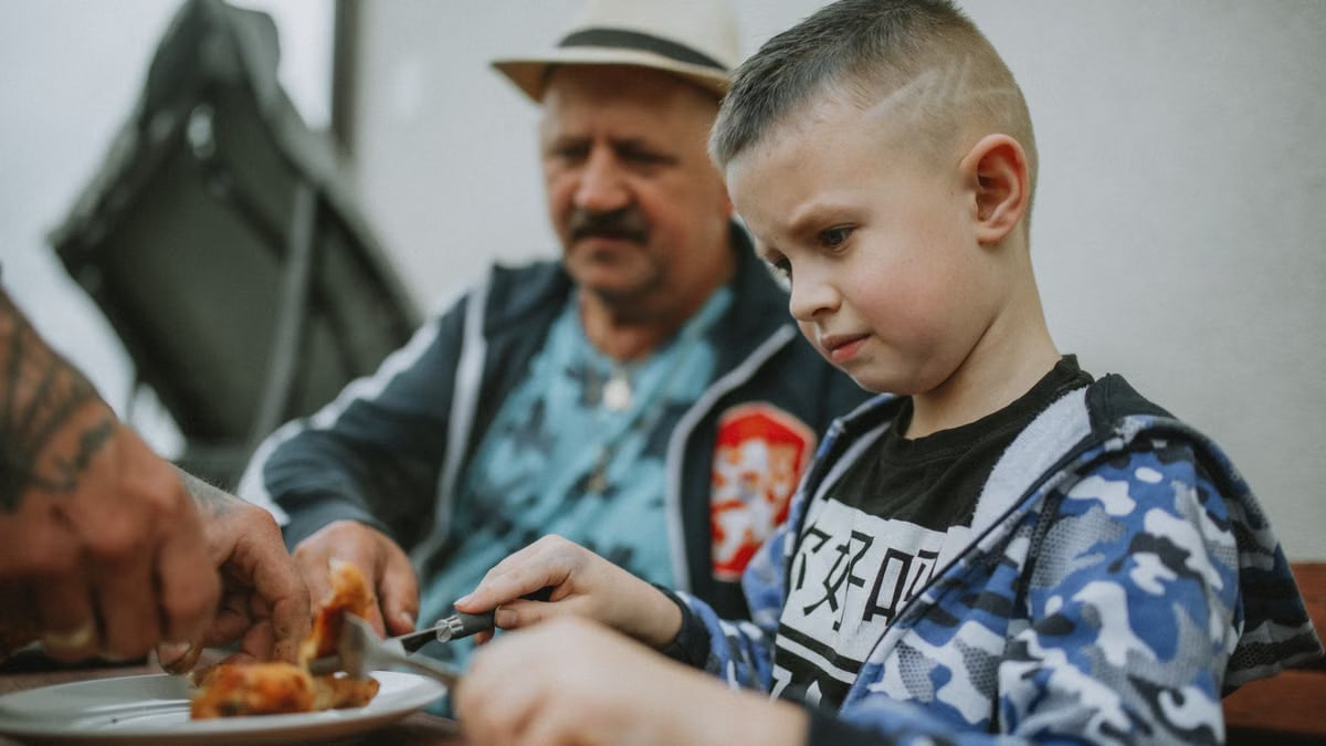 A child and his father share a meal outdoors, enjoying grilled food together.